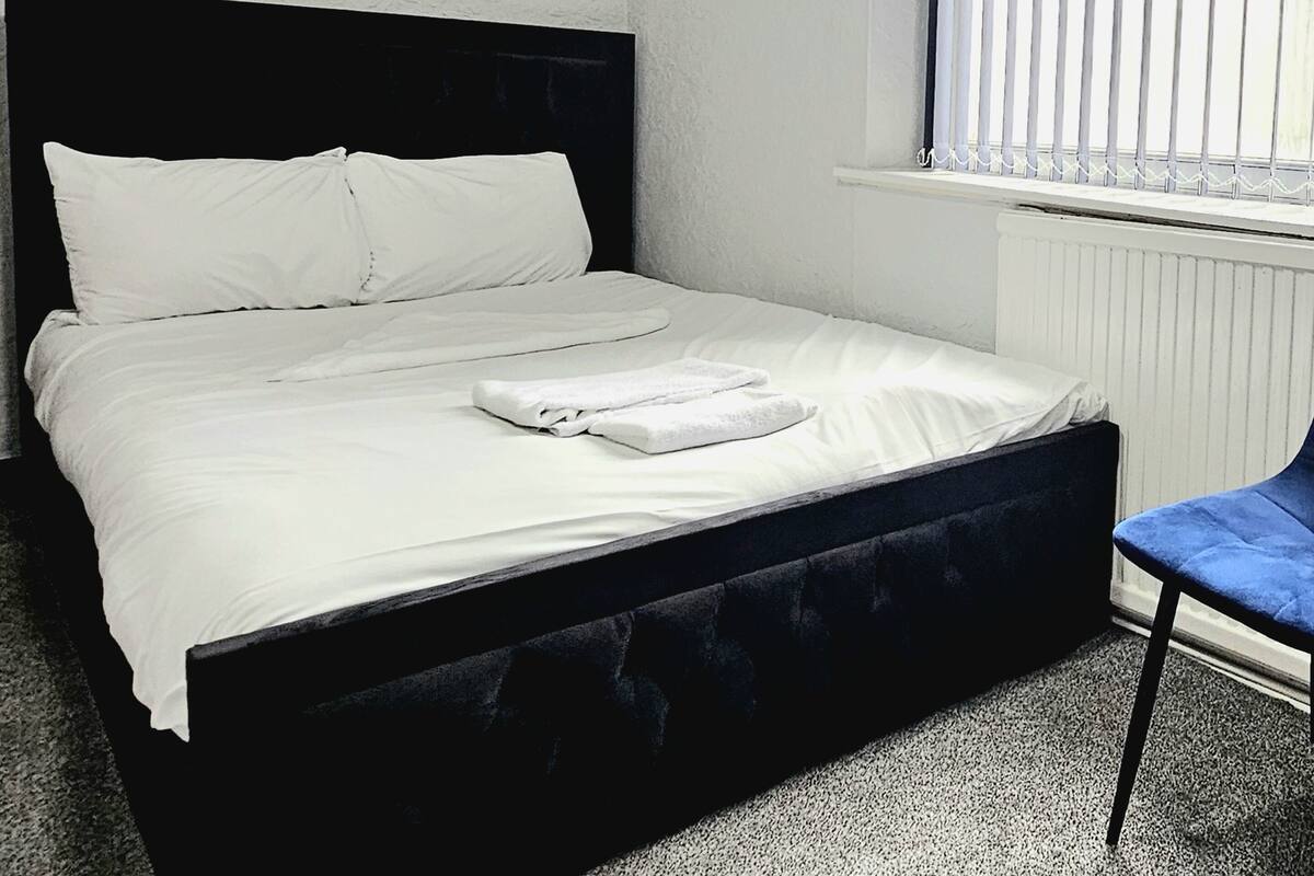 Una cama con ropa de cama blanca, una toalla doblada, un cabecero negro y una ventana con persianas.
