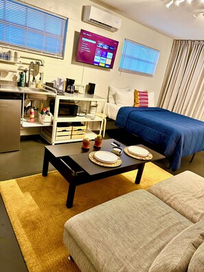 A compact hotel room with a kitchenette, a bed with a blue cover, a TV displaying a streaming service interface, and a window with blinds.