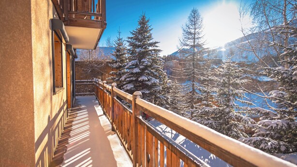 Un balcone con ringhiera in legno che si affaccia su un paesaggio innevato con alberi e montagne.