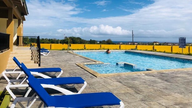 A pool area with blue lounge chairs, a clear sky, and a yellow wall in the background. - Royal Vacation Retreat (Falmouth)