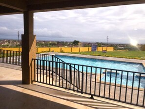 Piscina com guarda-corrimão de metal preto, cercada por uma vedação amarela, e vista para uma área residencial ao fundo.