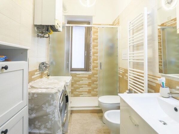 A modern bathroom with a washing machine, toilet, and a glass shower enclosure. - Primavera Guest House (Pontecagnano Faiano)