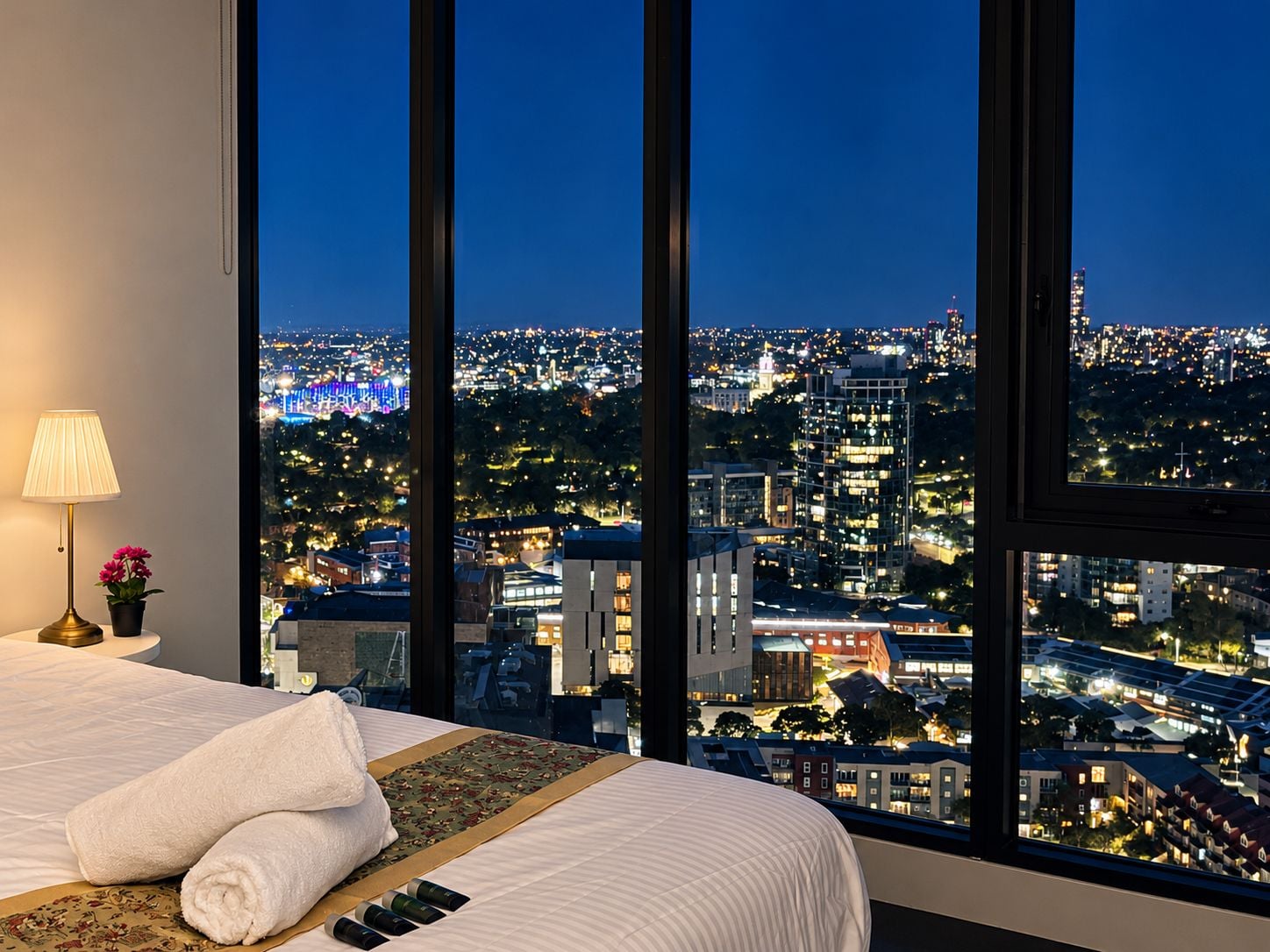 A hotel room with a large window offering a city view at night.