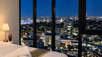 A hotel room with a large window offering a city view at night.