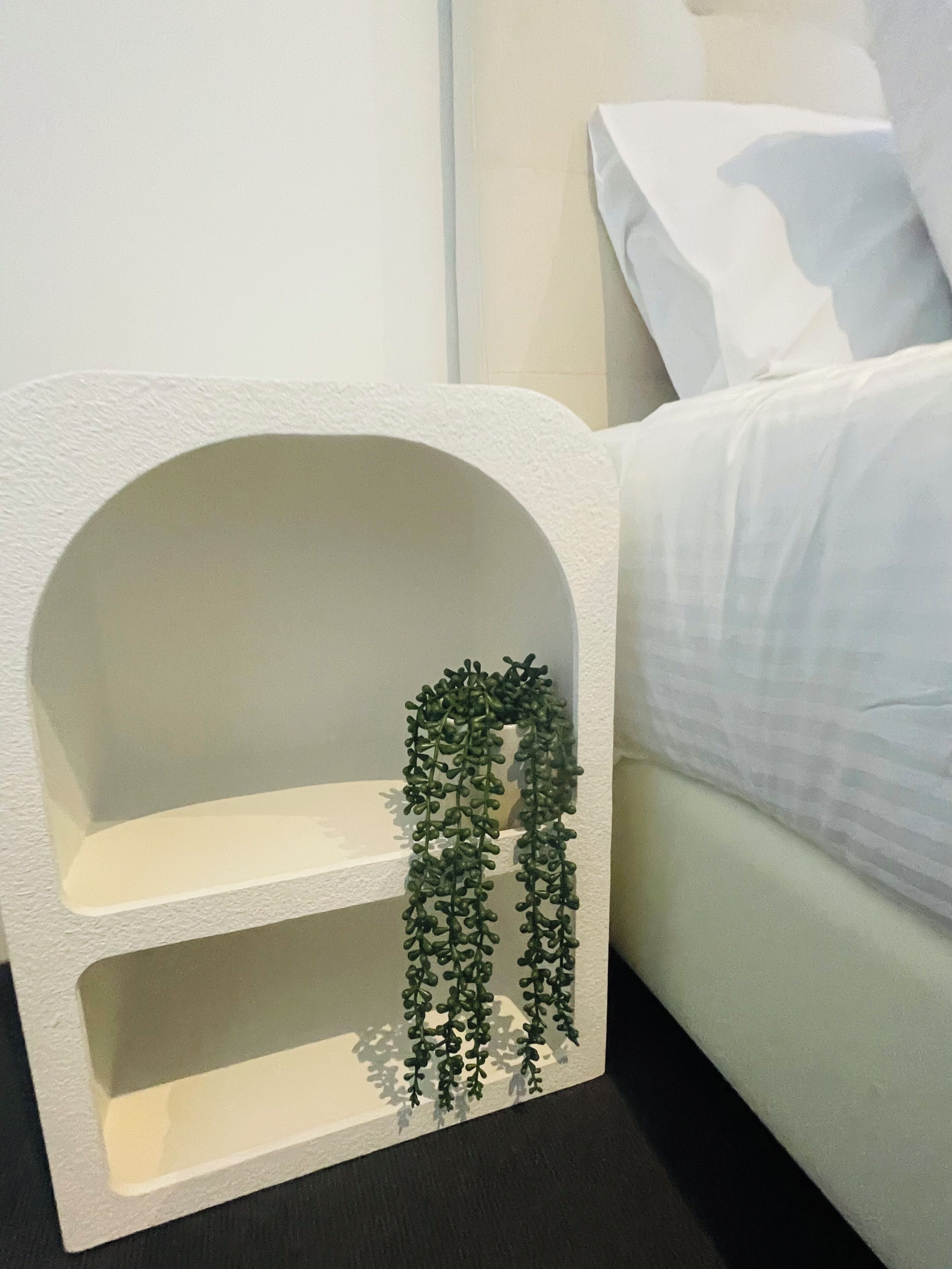 A white, arched, three-tiered shelf unit with hanging greenery against a wall, next to a bed with white bedding.