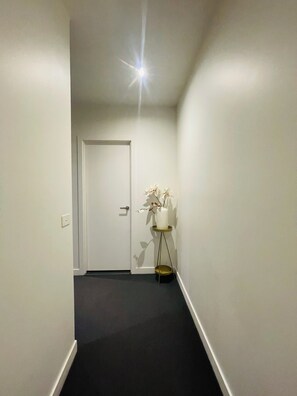 A narrow hallway with a white door at the end, a small potted plant on a gold stand, and a black carpet.