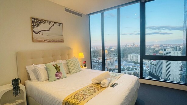 A hotel room with a large bed, a nightstand, a lamp, and a view of the cityscape through large windows.
