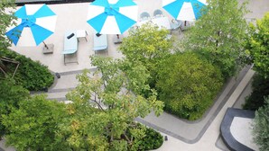 Vue aérienne d’un espace autour d’une piscine, avec des parasols bleus et blancs, des chaises longues et une végétation verdoyante.