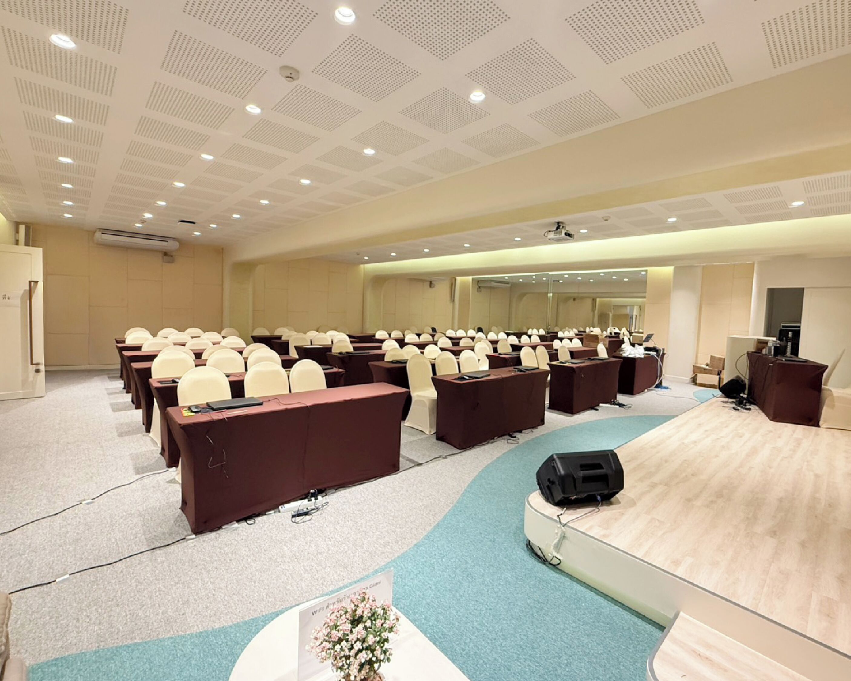 A conference room with rows of tables and chairs, a stage with a podium, and a large mirror on the back wall.