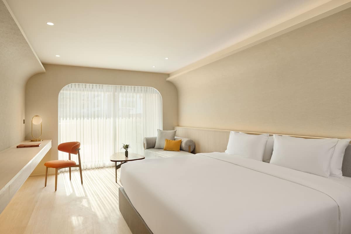 A modern hotel room with a large bed, a desk, a chair, and a small table with a lamp.