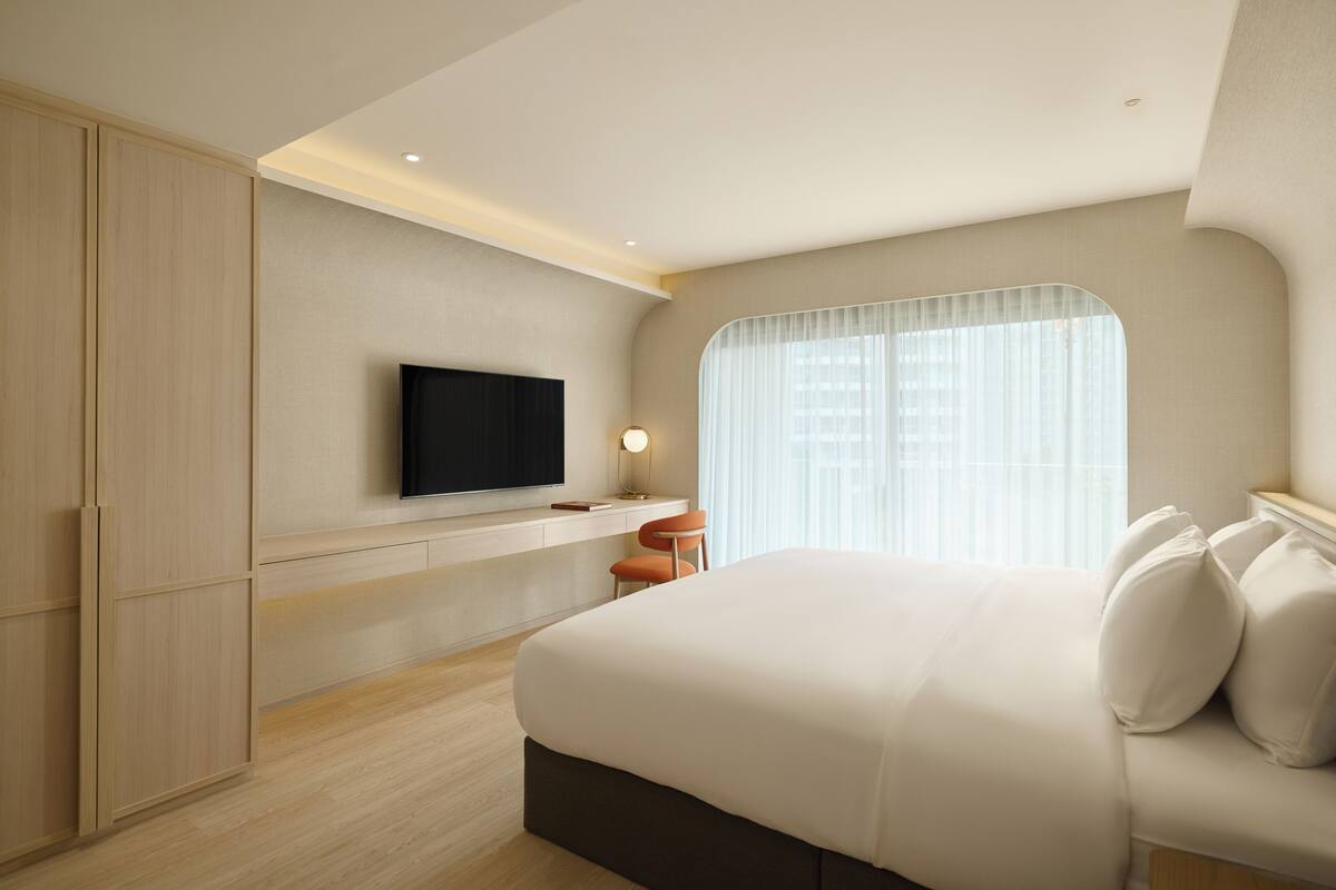 A modern hotel room with a large bed, a flat-screen TV mounted on the wall, a desk with a chair, and a window with sheer curtains.
