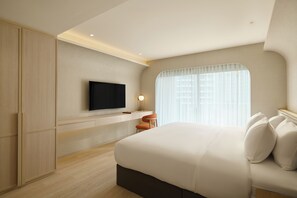 Una habitación de hotel moderna con una cama grande, un televisor de pantalla plana montado en la pared, un escritorio con una silla y una ventana con cortinas translúcidas.