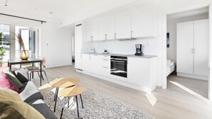 Une cuisine moderne avec des meubles blancs et un four noir, un coin repas avec une table et des chaises, et un salon avec un canapé et une table basse.