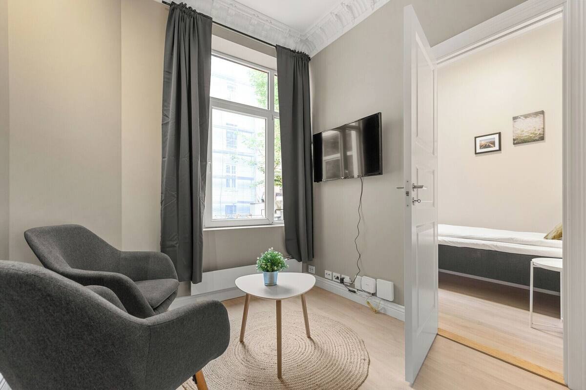 Une pièce moderne avec un canapé gris, une petite table ronde agrémentée d’une plante, un téléviseur fixé au mur et un lit avec un matelas et des oreillers.