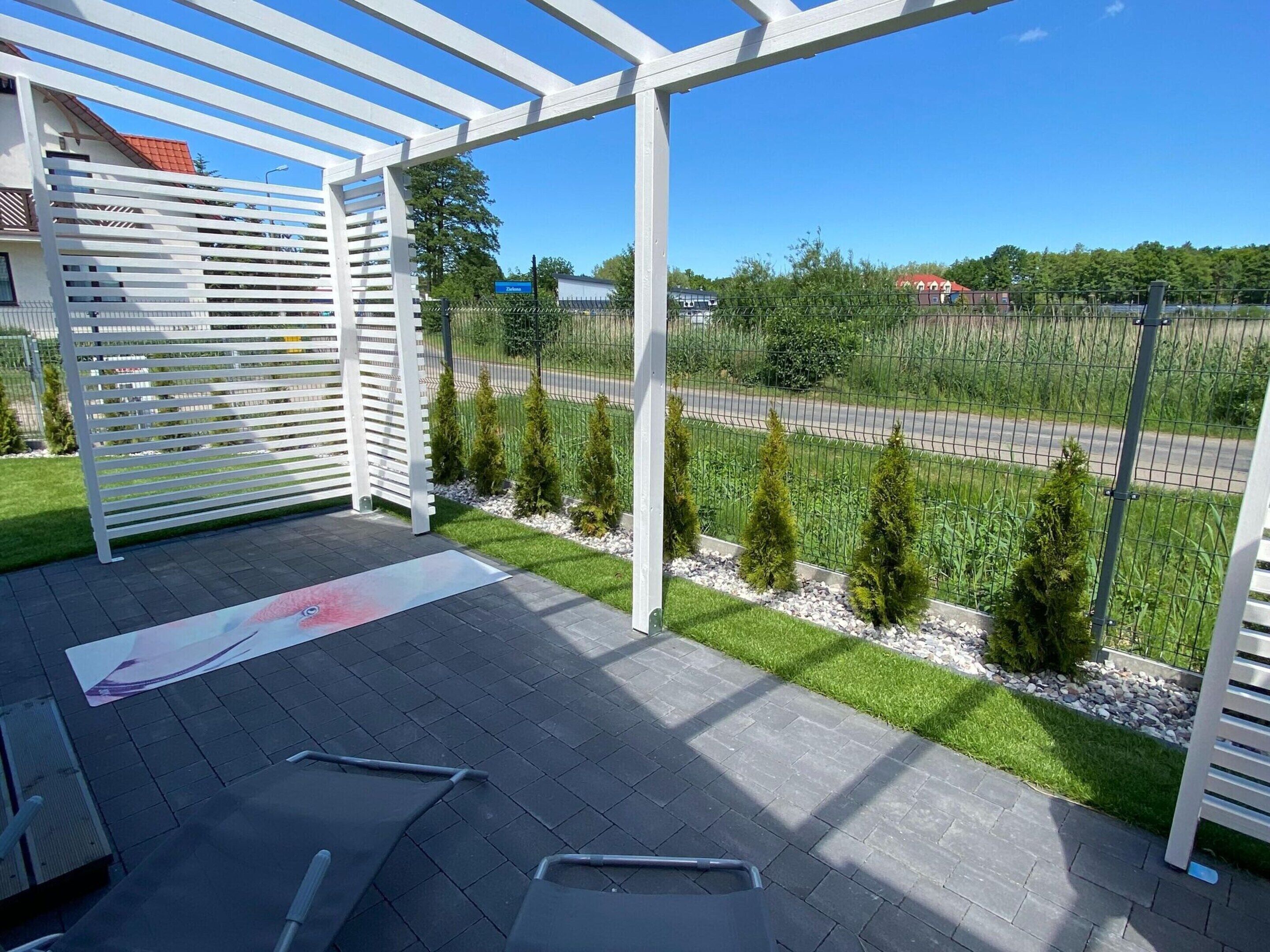 Un espacio exterior cubierto con una estructura de pérgola, un patio empedrado y vistas a una carretera y vegetación.