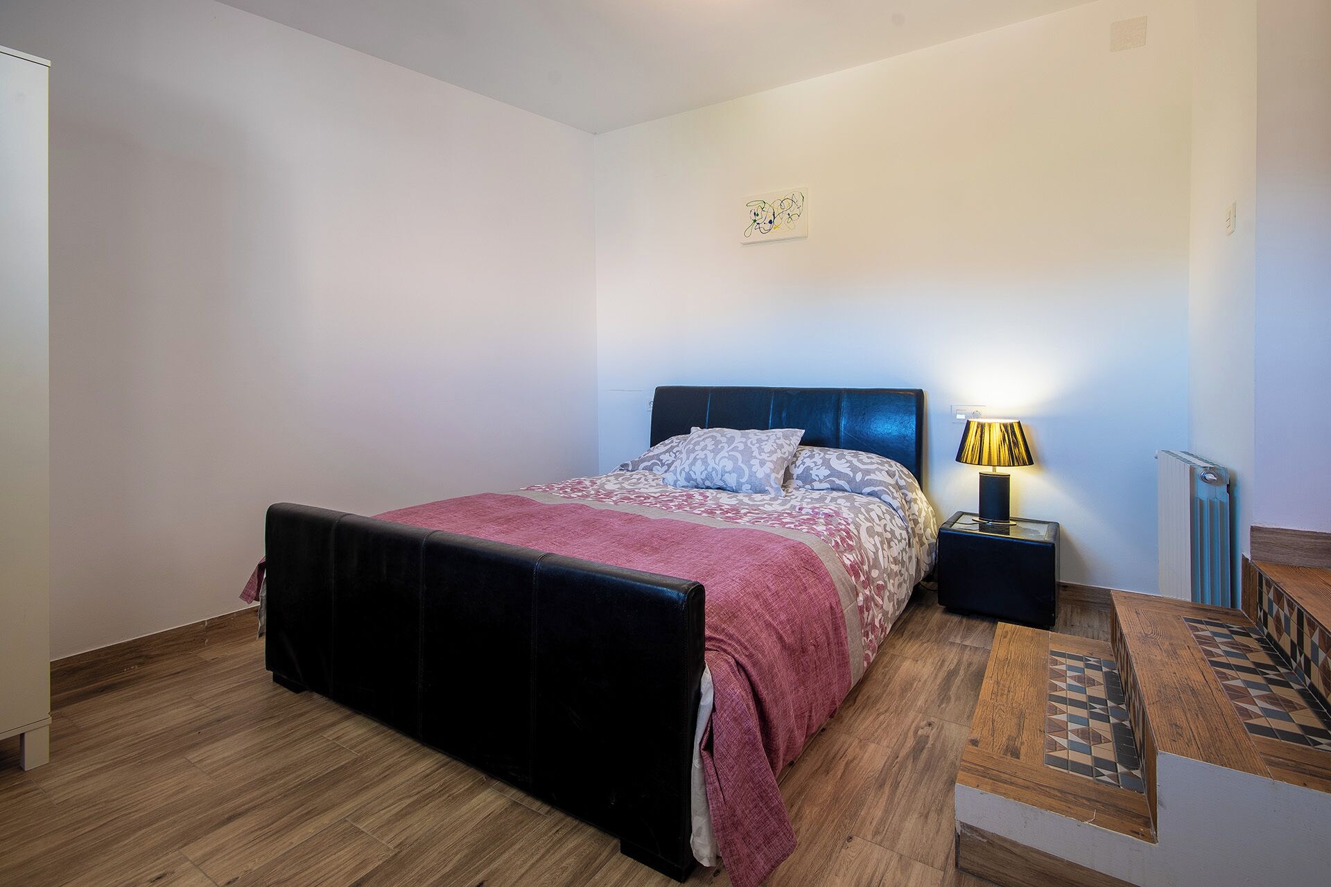 Un dormitorio con cama, mesita de noche con lámpara, radiador y una pequeña mesa de madera con camino de mesa estampado.