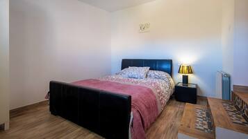 Un dormitorio con cama, mesita de noche con lámpara, radiador y una pequeña mesa de madera con camino de mesa estampado.