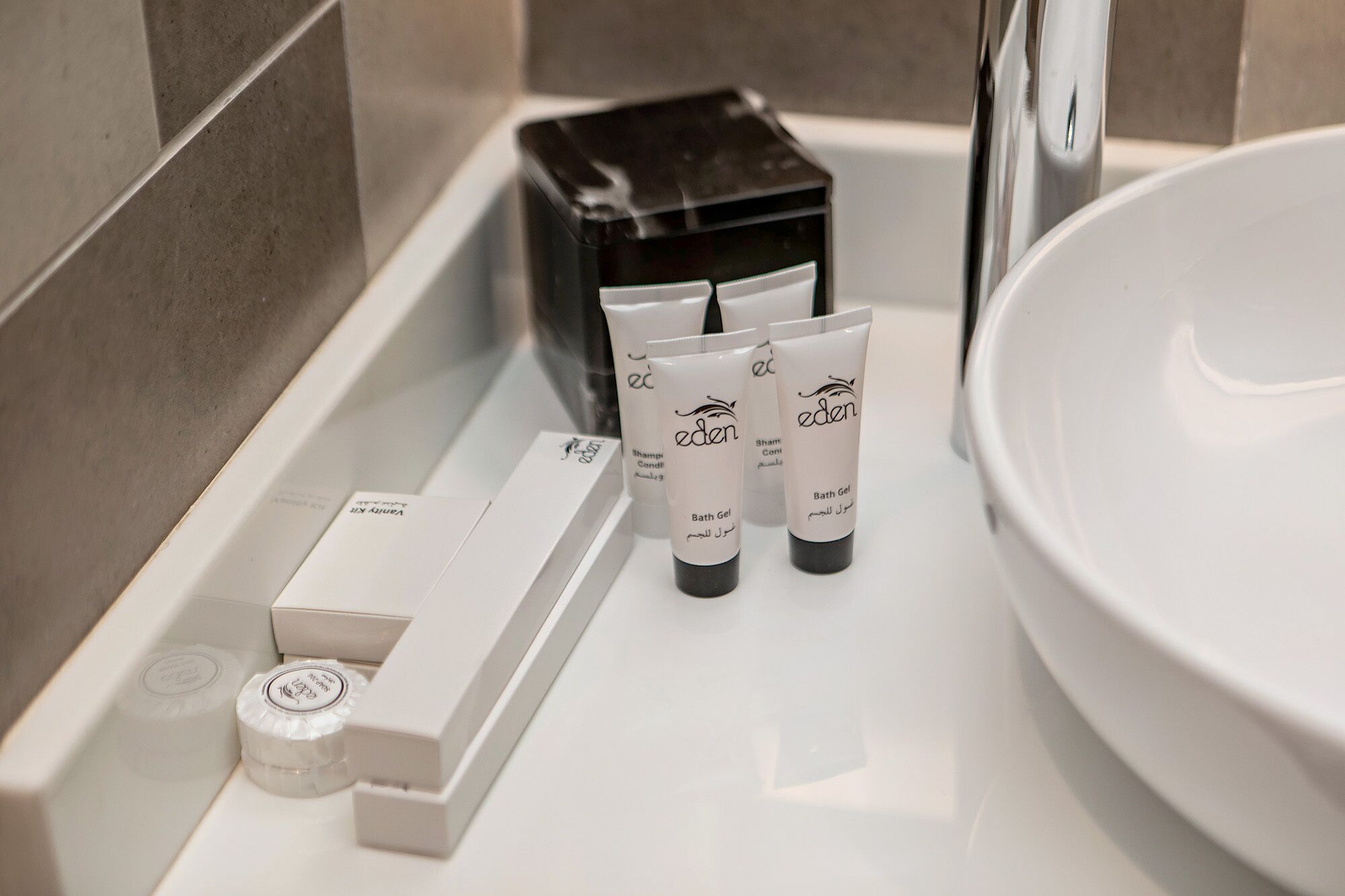 A bathroom with a white sink, three Eden bath gel tubes, a black soap dispenser, and a white bottle with a cap.