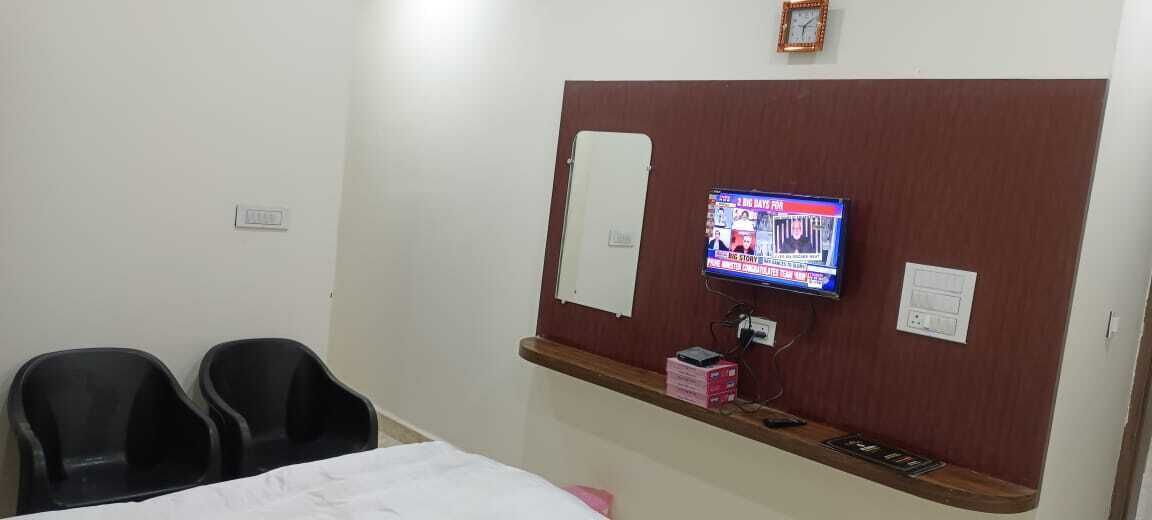 A hotel room with a bed, two black chairs, a TV mounted on the wall, a wooden shelf with items, and a clock.