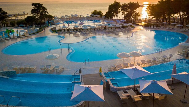 A large, multi-level swimming pool with sun loungers and umbrellas, surrounded by palm trees and a view of the ocean at sunset. - Zaton Resort Apts. With Pool (Nin)