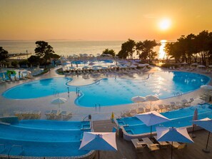 Una gran piscina de varios niveles con tumbonas y sombrillas, rodeada de palmeras y vista al océano al atardecer.