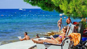 A beach scene with a wheelchair, a person reading, and others relaxing by the water. - Zaton Resort Apts. With Pool (Nin)
