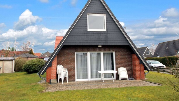 Ein zweistöckiges Haus mit dunklem Dach, einer Ziegel-Fassade und einer großen Schiebeglastür. Vor der Tür stehen zwei weiße Plastikstühle.