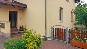 A two-story building with a beige exterior, a wooden gate, and a small garden area with various plants and flowers. - Behagliche Ferienwohnung im Idyllischen Korchow (Biendorf)