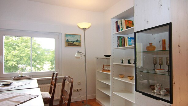 A room with a dining table, chairs, a floor lamp, a bookshelf, and a window with a view of trees. - Behagliche Ferienwohnung im Idyllischen Korchow (Biendorf)