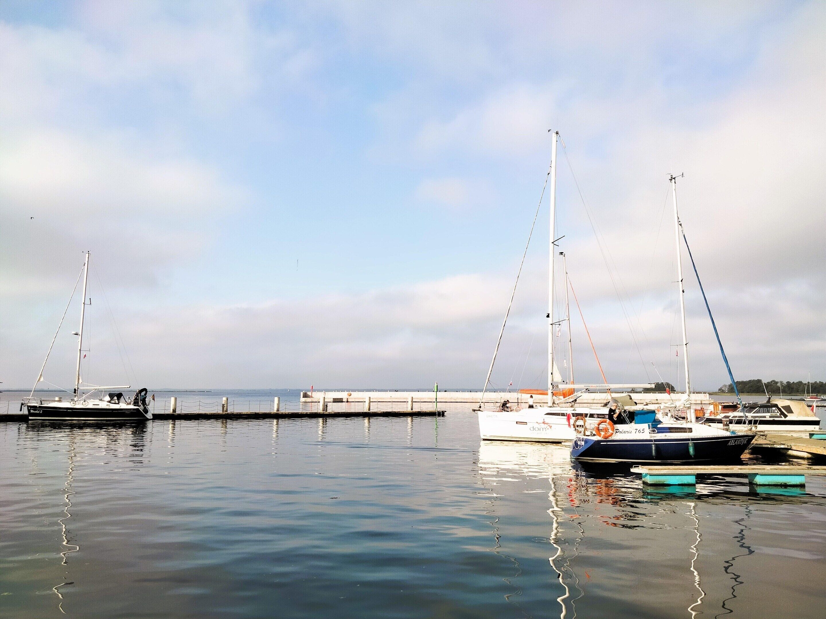 A marina with several sailboats docked, calm water, and a clear sky.