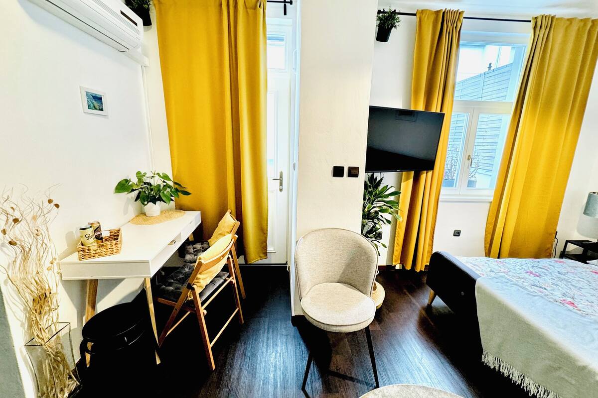 A room with a desk, chair, bed, TV, and yellow curtains.