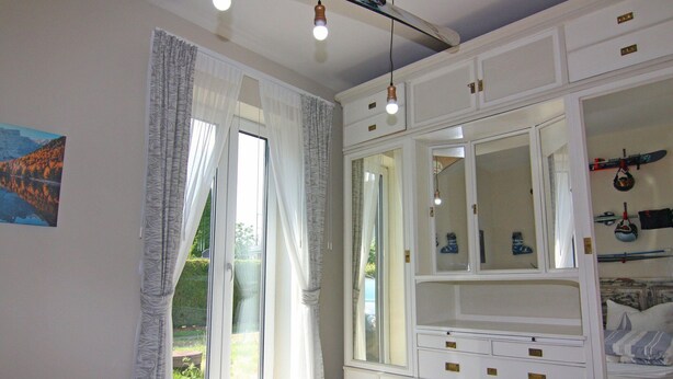 A bedroom with a bed, a large window with curtains, a wardrobe, and a mirror.