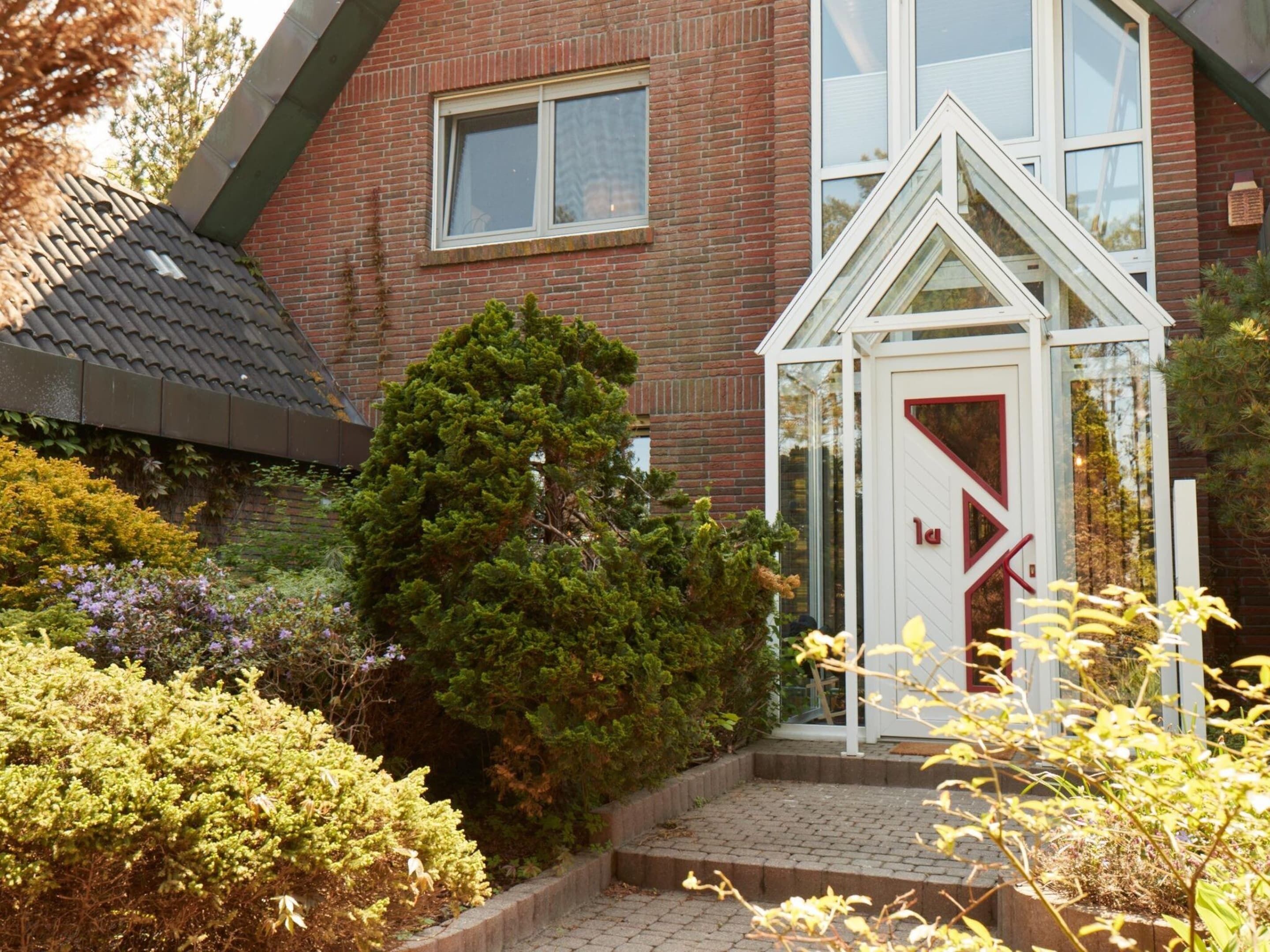 Une maison avec une façade en briques, un petit hall d’entrée vitré et un jardin bien entretenu, agrémenté de divers arbustes et d’un arbre.