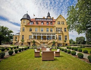 Un grand bâtiment jaune, doté d’un toit recouvert de tuiles rouges, d’un escalier central et d’une pelouse bien entretenue avec des bancs.
