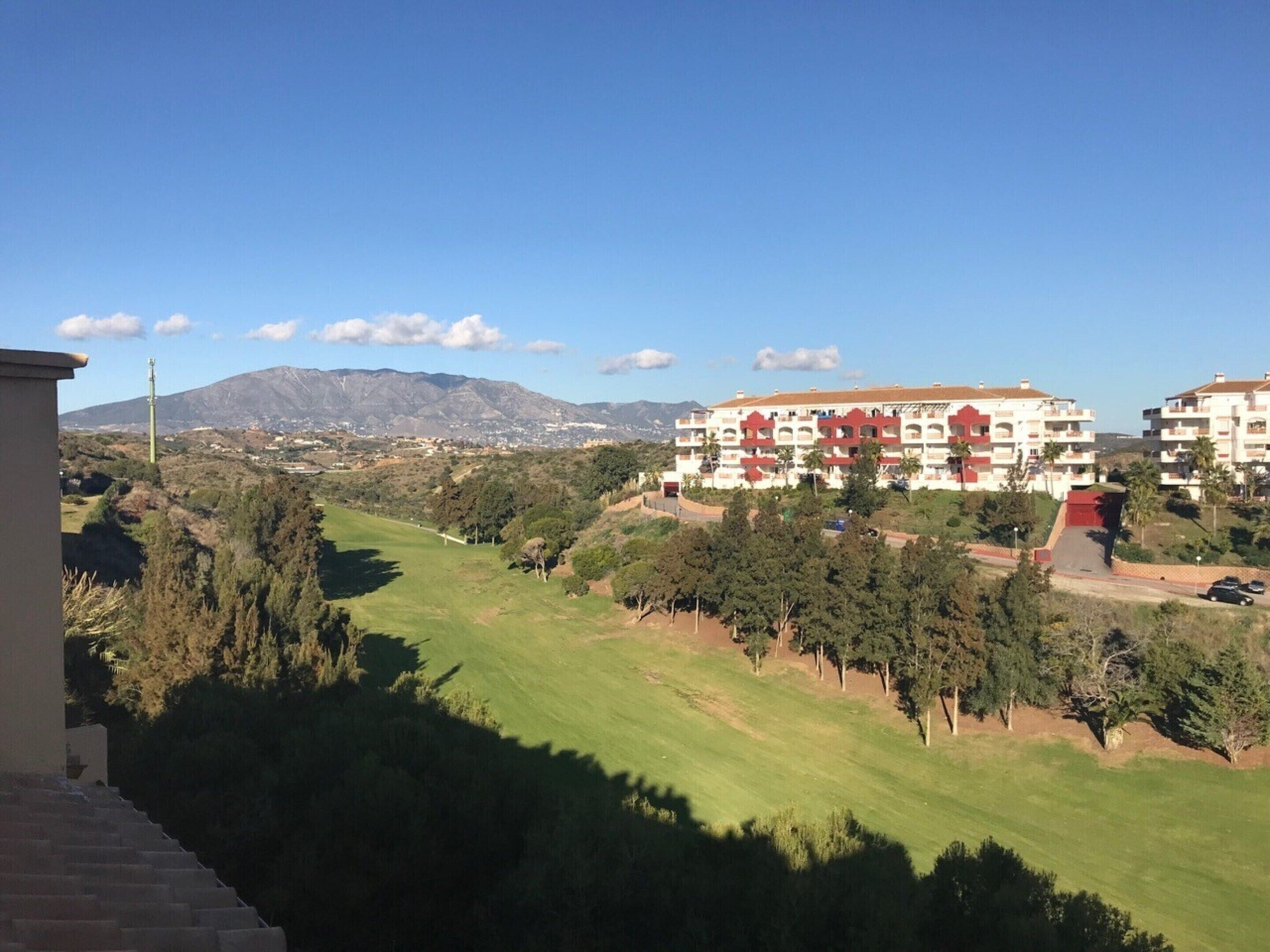 A residential area with a large building featuring balconies and a view of a green landscape with trees and a distant mountain.