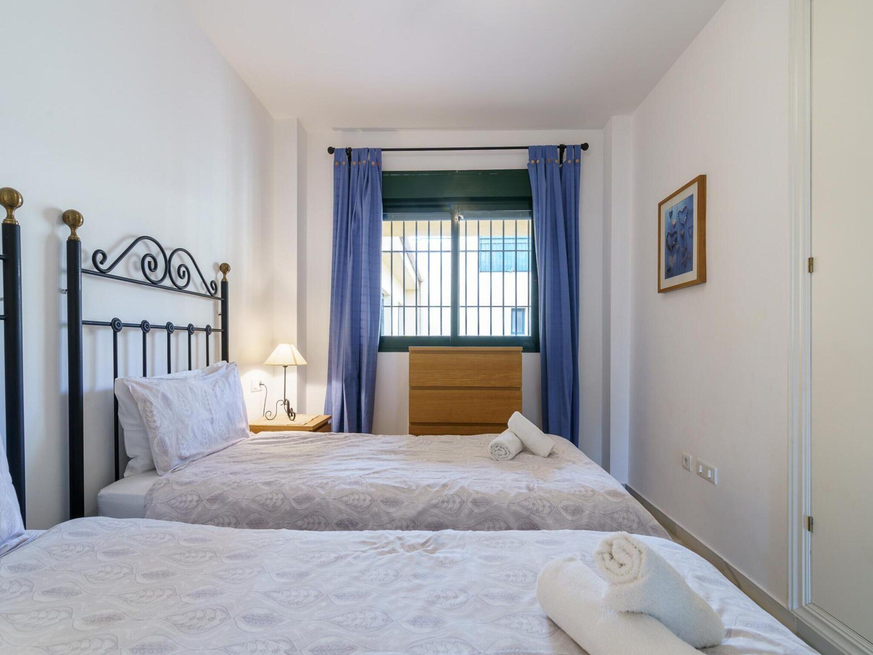 A bedroom with two beds, a window with blue curtains, a bedside table with a lamp, and a framed picture on the wall.