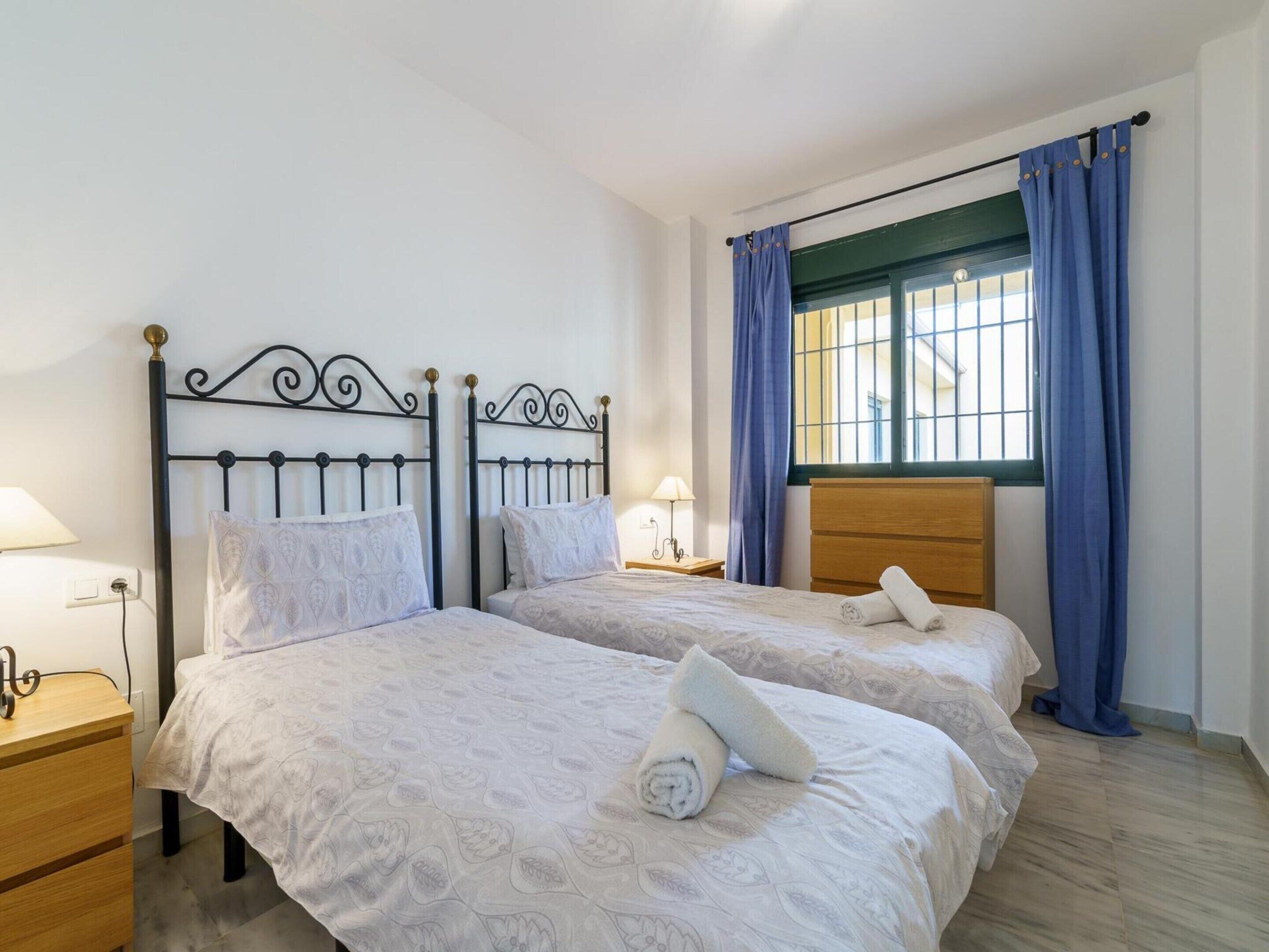 A hotel room with two beds, a wooden nightstand, a lamp, and a window with blue curtains.