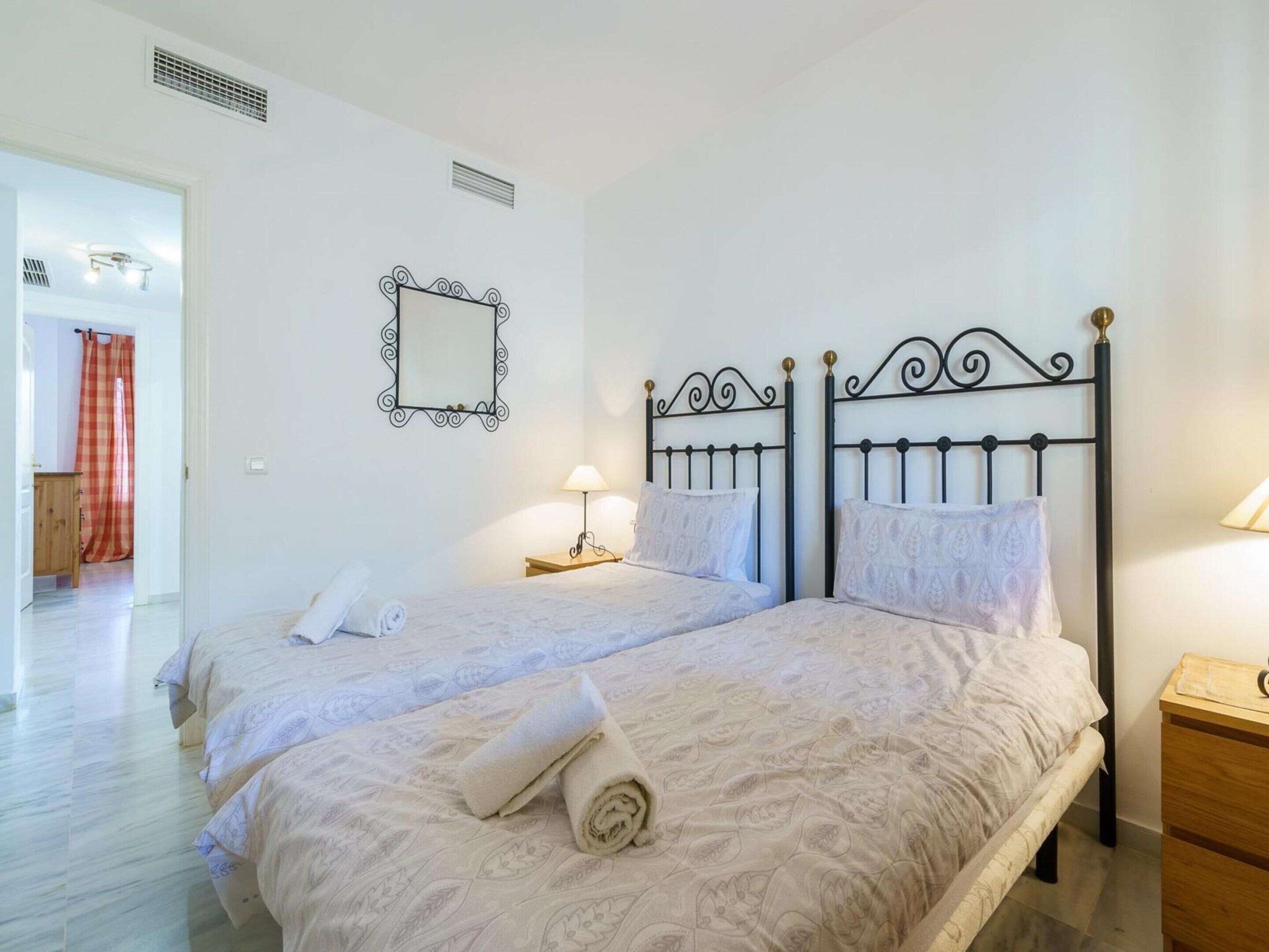 A double bed with a patterned bedspread, two bedside tables with lamps, a mirror on the wall, and a wooden door with a red curtain.