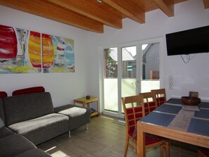 A living room with a grey sofa, wooden dining table with chairs, a small side table, and a TV mounted on the wall. - Doppelhaushalfte Luv in Zingst (Zingst)