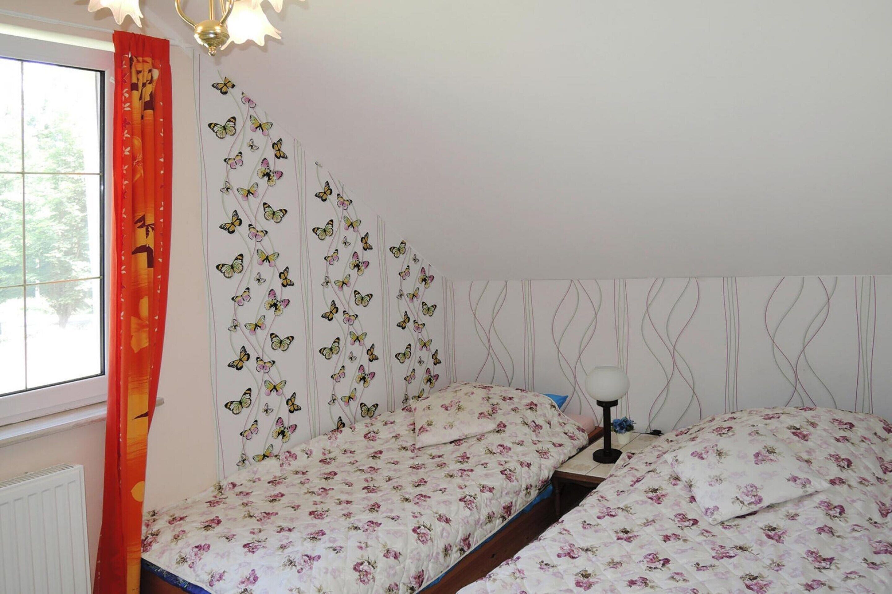 A bedroom with two beds, butterfly wall art, a window with curtains, and a bedside lamp.