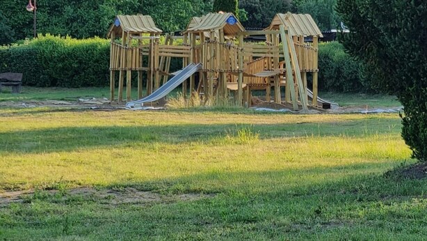 Ein Spielplatz mit verschiedenen Holzspielgeräten, darunter eine Rutsche und Klettergerüste, inmitten einer Wiese mit Bäumen und einem Gebäude im Hintergrund.