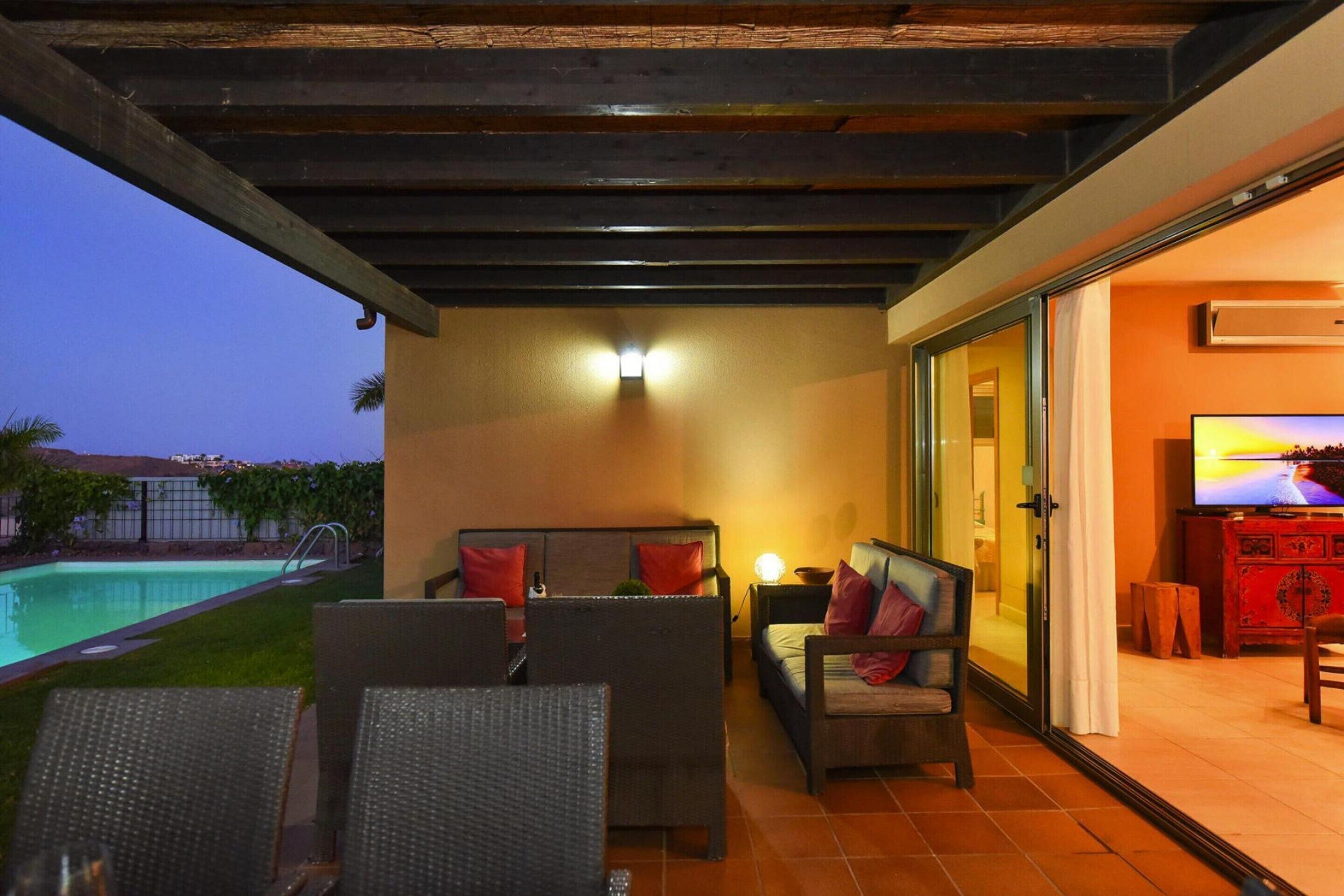 A covered outdoor seating area with wicker furniture, a pool, and a television in the background.