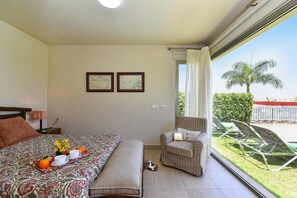 A hotel room with a bed, a chair, a small table with a tray of fruit and cups, and a window with a view of palm trees and a fence. - Holiday Home in Maspalomas (San Bartolomé de Tirajana)