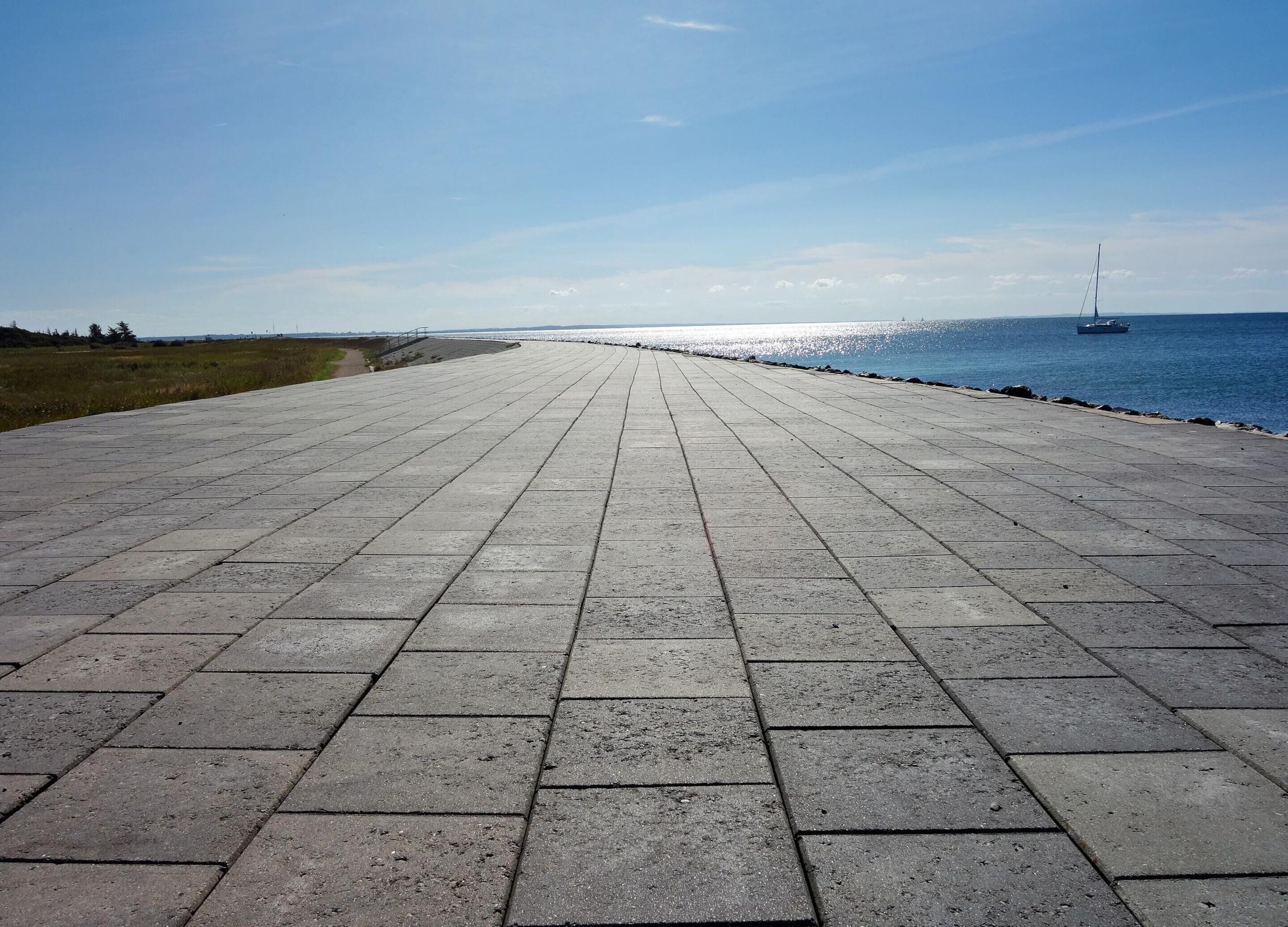 A paved walkway by the sea with a clear view of the ocean and a sailboat in the distance.