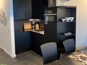 A compact kitchen with dark cabinetry, a coffee maker, and a small dining area with a checkered tablecloth. - Ferienwohnung mit Meerblick in Wyk auf Foehr (Wyk auf Foehr)