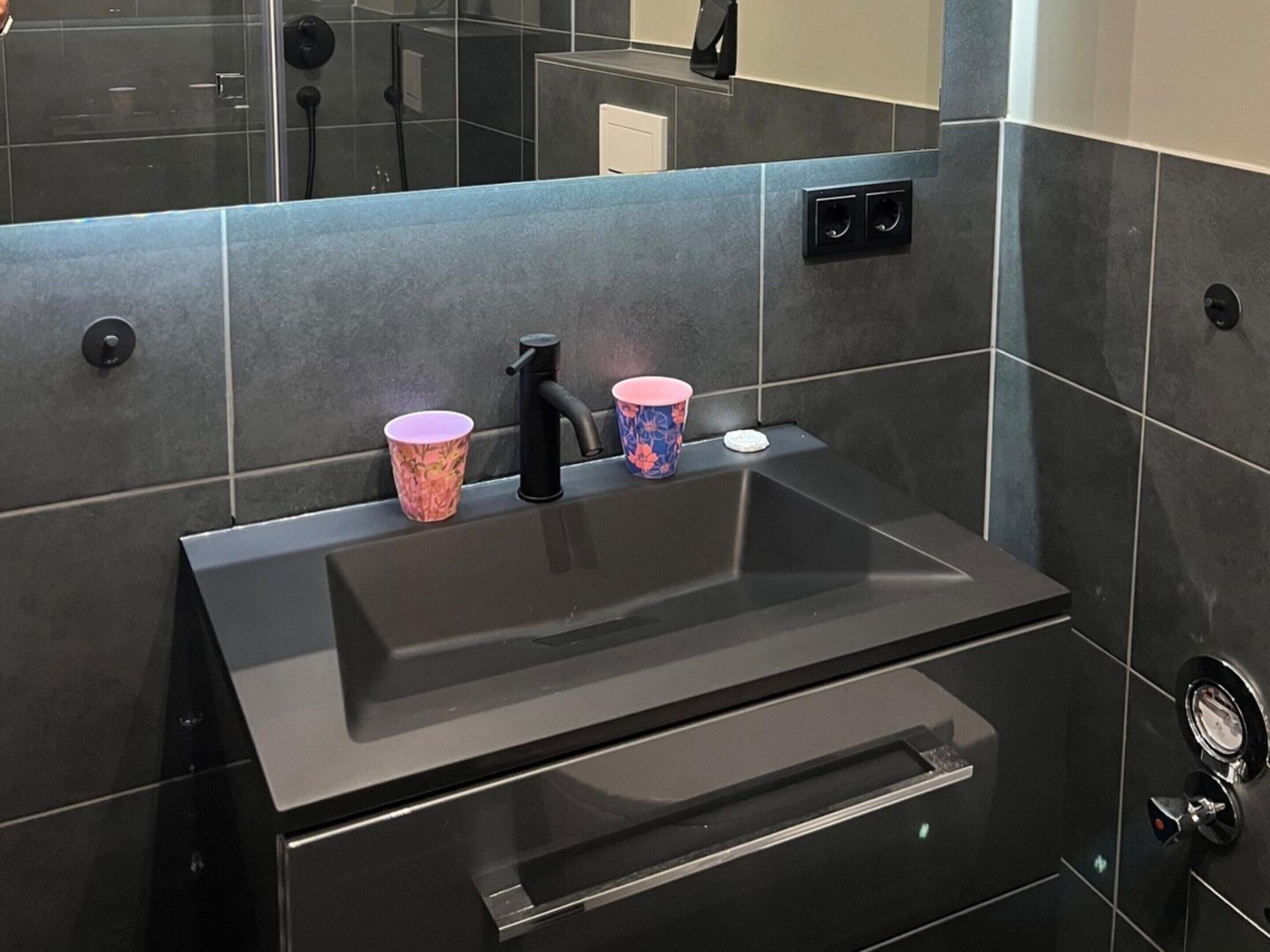 Un baño moderno con un lavamanos negro, dos tazas y un espejo.