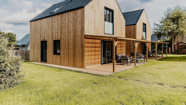 Une maison moderne en bois, dotée d’un toit à pente, d’un patio couvert avec des meubles d’extérieur et d’une pelouse bien entretenue.