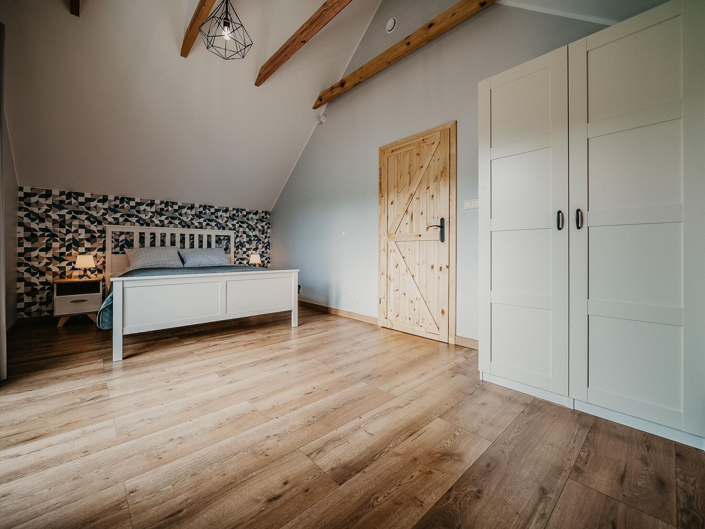 Un dormitorio con techo inclinado, cama con cabecera de estampado geométrico, armario de madera y una puerta de madera grande.