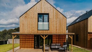 Une maison moderne en bois, dotée d’une grande baie vitrée, d’un patio couvert avec des meubles d’extérieur et d’une pelouse bien entretenue.