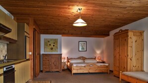 A cozy room with a wooden ceiling, a bed with a wooden headboard, a nightstand with a lamp, a wooden wardrobe, and a kitchen area with a stove and cabinets.